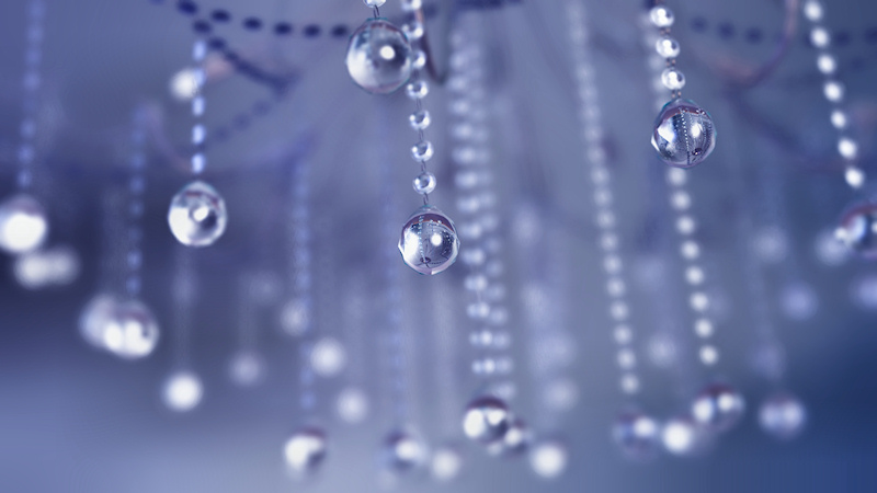 A 3D macro shot of a crystal chandelier. Myriads of strands of sparkling glass beads catch the light to create a lovely bokeh in the background. The blurred glass drops are reflected clearly in the sharply focused crystal balls in the foreground. The silver branches of the candelabra and soft blue atmosphere and lights give an early evening glow to the piece.
